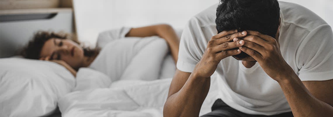 A man holds his face while a woman sleeps in a bed behind him