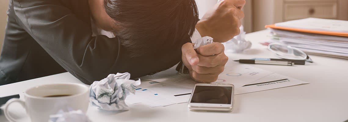 Frustrated man with his head on a desk surrounded by crumpled paper